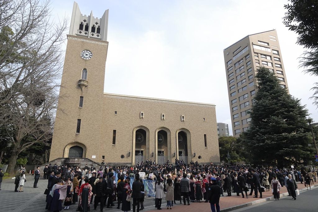  Large group of people gathered in front of Okuma Auditorium at Waseda University, with a tall beige clock tower and modern high-rise in the background.