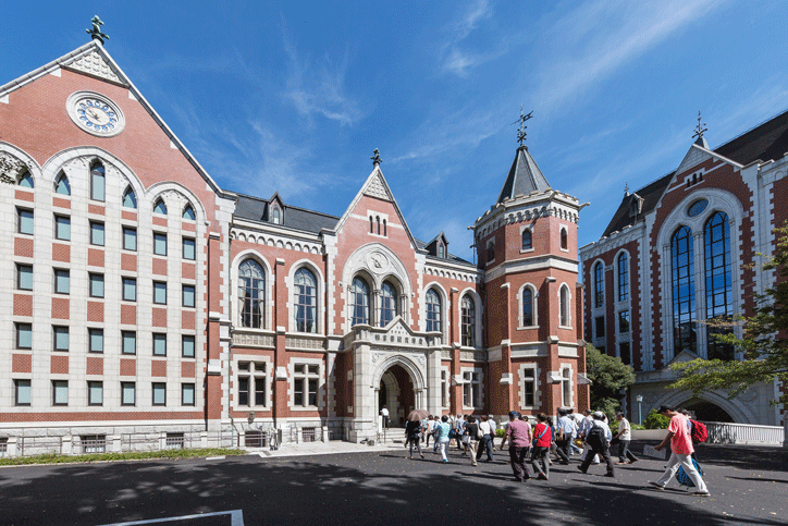 Group of people walking in front of the red-brick Gothic Revival-style Mita Campus building at Keio University, under a clear blue sky.