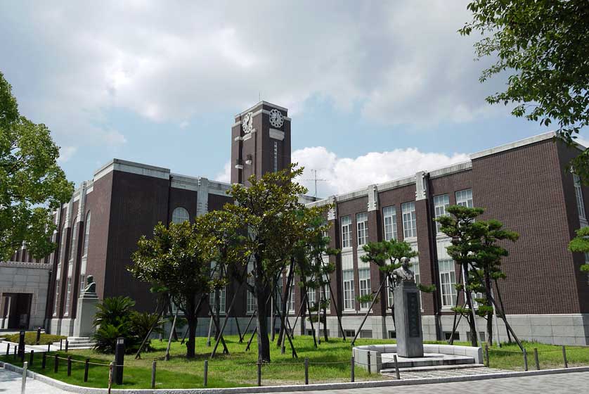 A view of Kyoto University's Clock Tower Centennial Hall, featuring brown brick architecture, traditional pine trees, and a clear sky.