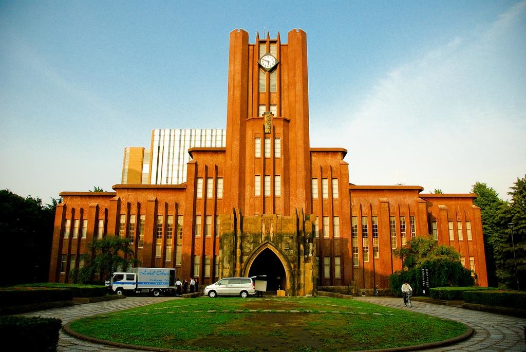 Historic red-brick building of the University of Tokyo with a central clock tower, surrounded by green lawns and a few parked vehicles.